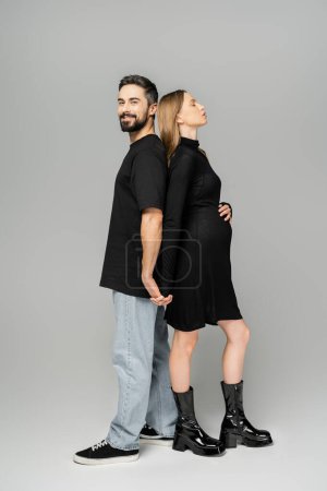 Photo for Full length of smiling bearded man looking at camera while holding hand and standing back to back with stylish pregnant wife in black dress on grey background, new beginnings and parenting concept - Royalty Free Image
