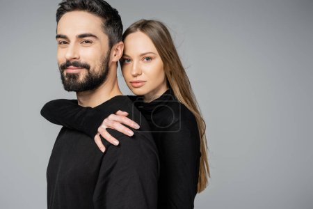Retrato de mujer de pelo justo con maquillaje natural abrazando sonriente y barbudo marido mientras mira a la cámara aislada en gris, marido y mujer concepto de relación