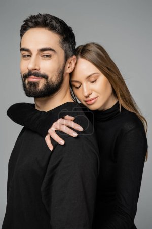 Porträt einer entspannten blonden Frau in schwarzer Kleidung, die einen bärtigen Ehemann umarmt, während sie isoliert auf einem grauen Beziehungskonzept zwischen Mann und Frau steht
