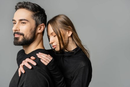 Mujer cabellera en ropa negra abrazando sonriente y barbudo marido en camiseta mientras que de pie juntos aislados en gris, marido y mujer concepto de relación