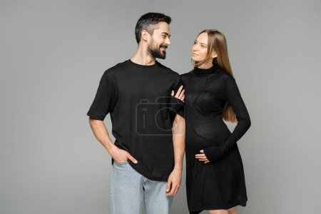 Mujer embarazada positiva en vestido negro tocando brazo de marido barbudo alegre en camiseta y jeans mientras está de pie aislado en gris, nuevos comienzos y el concepto de crianza  