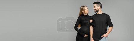 Stylish and pregnant woman in black dress touching arm of smiling bearded husband and standing together isolated on grey, new beginnings and parenting concept, banner 