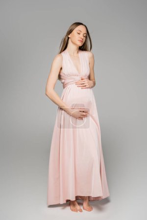 Longitud completa de mujer embarazada descalza y de pelo justo en vestido rosa elegante tocando el vientre y de pie con los ojos cerrados sobre fondo gris, traje de embarazo elegante y elegante