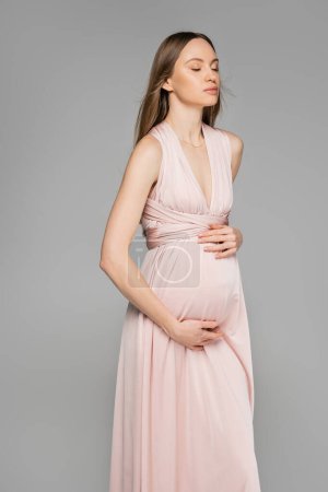 Modische hellhaarige und schwangere Frau in rosa Kleid, die Bauch berührt und mit geschlossenen Augen auf grauer, eleganter und stilvoller Schwangerschaftskleidung steht, Sinnlichkeit, werdende Mutter 