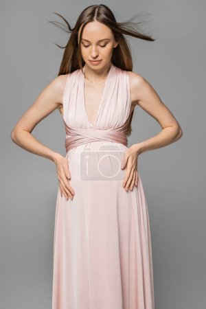 Fashionable and long haired pregnant woman in pink dress touching belly and looking down while standing isolated on grey, elegant and stylish pregnancy attire, sensuality, mother-to-be 