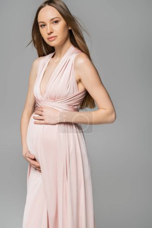 Femme enceinte aux cheveux blonds et à la mode en robe rose regardant la caméra debout et posant isolée sur des vêtements de grossesse gris, élégants et élégants, sensualité, future mère 