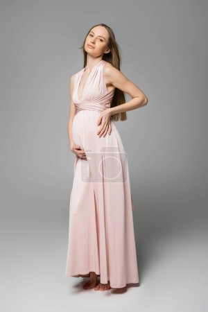 Trendige und hellhaarige schwangere Frau in rosa Kleid, die in die Kamera schaut, während sie auf grauem Hintergrund posiert, elegante und stilvolle Schwangerschaftskleidung, Sinnlichkeit, werdende Mutter 