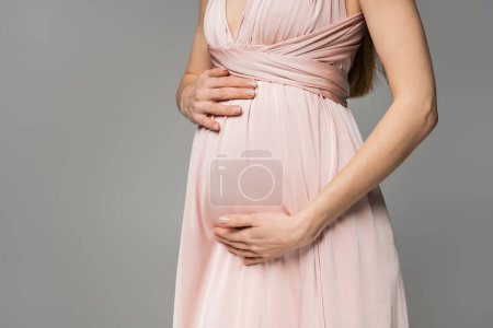 Ausgeschnittene Ansicht einer modischen und langhaarigen Frau in rosa Kleid, die den Bauch berührt, während sie isoliert auf grauer, eleganter und stilvoller Schwangerschaftskleidung steht, Sinnlichkeit, werdende Mutter 