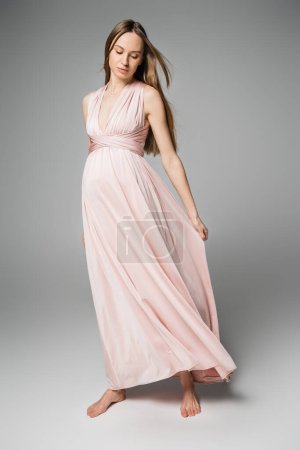 Madre embarazada descalza y de moda en vestido rosa posando y tocando tela sobre fondo gris, elegante y elegante atuendo de embarazo, sensualidad, futura madre 
