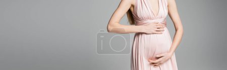 Vista recortada de la mujer de moda y embarazada en vestido rosa tocando el vientre mientras está de pie aislado en gris con espacio para copiar, elegante y elegante atuendo de embarazo, pancarta 