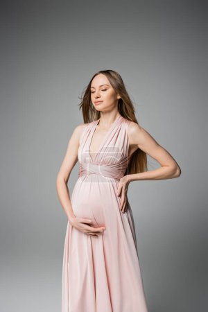 Fashionable long haired and pregnant woman with closed eyes touching belly while posing in pink dress isolated on grey, elegant and stylish pregnancy attire, sensuality, mother-to-be 