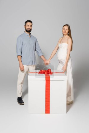 Smiling pregnant woman holding hand of husband and looking at camera while standing near big git box during gender reveal surprise party on grey background, expecting parents concept