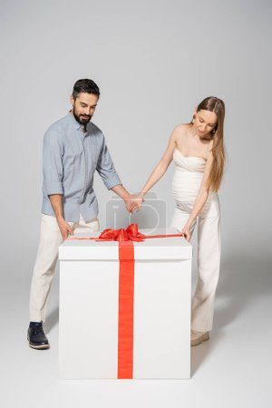 Full length of stylish couple holding hands while standing together near bog gift box during gender reveal surprise party and celebration on grey background, expecting parents concept