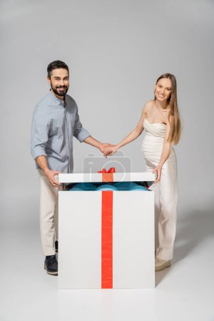 Cheerful and stylish couple holding hands while opening bog gift box with blue balloons during gender reveal surprise party on grey background, expecting parents concept, it`s a boy