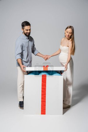 Positive man holding hand of pregnant wife while opening gift box with blue balloons during gender reveal surprise party on grey background, expecting parents concept, it`s a boy 
