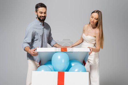 Cheerful and trendy expecting parents opening big gift box with festive blue balloons during gender reveal surprise party and celebration isolated on grey, it`s a boy