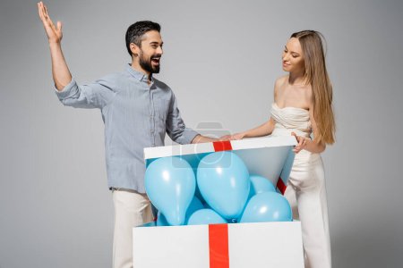 Excited bearded man looing at stylish pregnant wife opening big gift box with blue balloons during gender reveal surprise party and celebration isolated on grey, expecting parents concept, it`s a boy 