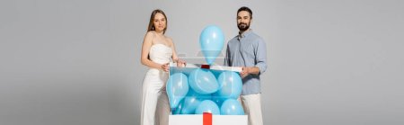 Smiling and trendy expecting parents looking at camera while opening bog gift box with festive blue balloons during gender reveal surprise party isolated on grey, it`s a boy, banner