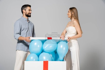 Cheerful and stylish pregnant woman looking at husband while opening big gift box with festive blue balloons during gender reveal surprise party isolated on grey, expecting parents, it`s a boy