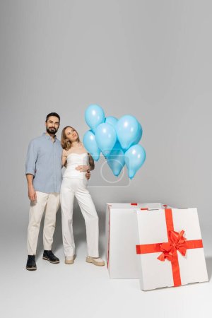 In voller Länge lächelnde und stylische werdende Eltern, die sich umarmen, während sie in der Nähe einer großen Geschenkbox mit festlich blauen Luftballons stehen, während die Geschlechterparty auf grauem Hintergrund eine Überraschung enthüllt, es ist ein Junge 
