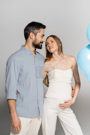 Lächelnder und stylischer bärtiger Mann, der schwangere Frau umarmt und ansieht, während er beim Geschlechtsverkehr neben blauen Festballons steht, verrät Überraschungsparty auf grauem Hintergrund, erwartete Eltern