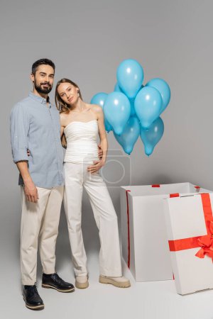Voller positiver werdender Eltern, die sich umarmen und in die Kamera schauen, in der Nähe einer großen Geschenkbox mit festlich blauen Luftballons während der Geschlechterparty auf grauem Hintergrund, es ist ein Junge 