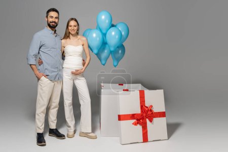 Volle Länge von fröhlichen und trendigen werdenden Eltern, die sich umarmen und in die Kamera schauen, während sie in der Nähe einer großen Geschenkbox stehen und festliche blaue Luftballons während des Geschlechtsverkehrs verraten Überraschungsparty auf grauem Hintergrund