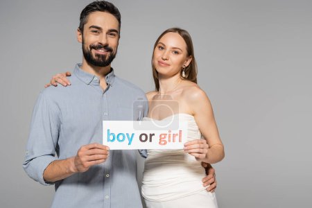 Photo for Positive and trendy pregnant woman holding card with boy or girl lettering and hugging stylish husband during gender reveal surprise party isolated on grey, expecting parents concept - Royalty Free Image