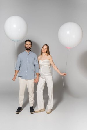 Full length of positive and stylish expecting parents looking at camera while holding hands and balloons during gender reveal surprise party on grey background, expecting parents concept