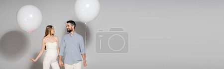 Fashionable pregnant woman holding hand of smiling husband and festive balloon during gender reveal surprise party on grey background with copy space, banner, expecting parents concept