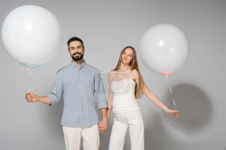 Lächelnde und stylische werdende Eltern, die Händchen halten und festlich weiße Luftballons, während sie beim Geschlechtsverkehr in die Kamera schauen, verraten Überraschungsparty auf grauem Hintergrund