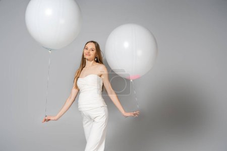 Positive und modische schwangere Frau, die in die Kamera schaut, während sie weiße festliche Luftballons während des Geschlechtsverkehrs hält, verrät Überraschungsparty auf grauem Hintergrund, modische Schwangerschaftskleidung
