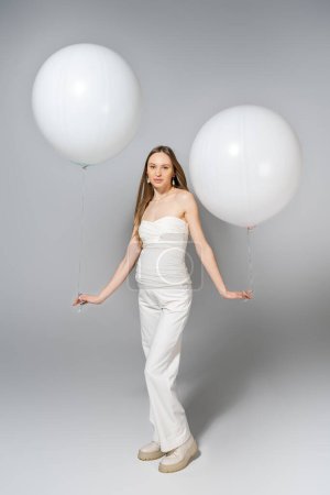 Volle Länge der trendigen und hellhaarige schwangere Frau mit weißen Luftballons und Blick in die Kamera während des Geschlechts offenbaren Überraschungsparty auf grauem Hintergrund, modische Schwangerschaftskleidung