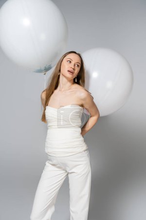 Modische schwangere Frau, die wegschaut, während sie festliche weiße Luftballons in der Hand hält und beim Geschlechtsverkehr steht, verrät Überraschungsparty auf grauem Hintergrund, modische Schwangerschaftskleidung