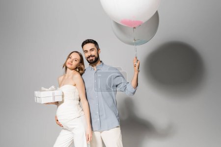 Positive and stylish man holding festive balloons and hugging pregnant wife with gift box during celebration and gender reveal surprise party on grey background, pregnancy attire, boy or girl 