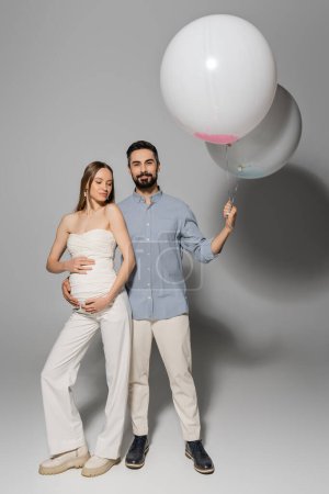 Volle Länge der positive und stilvolle Mann hält festliche Luftballons und umarmt schwangere Frau während Baby-Dusche Party auf grauem Hintergrund, erwartete Eltern Konzept, Junge oder Mädchen 