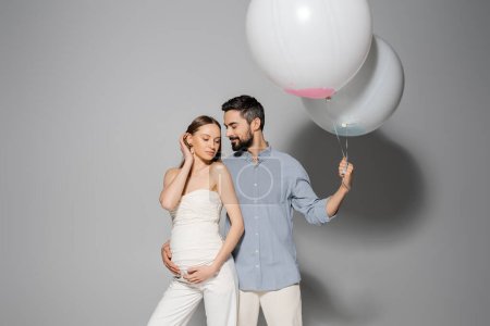 Lächelnder und bärtiger Mann umarmt stilvolle schwangere Frau und hält festliche Luftballons während Geschlechterparty und Feier auf grauem Hintergrund, werdende Eltern, Junge oder Mädchen 