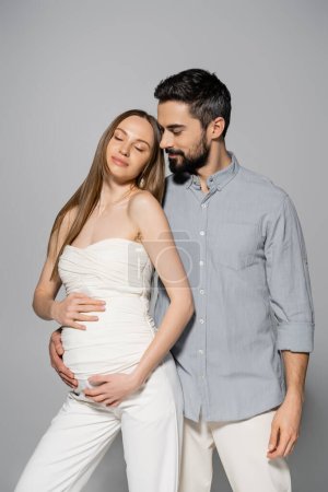 Bärtiger und stilvoller Mann umarmt entspannte und modische schwangere Frau, während er auf grauem Hintergrund zusammen steht, werdende Eltern, Babybauch, Mann und Frau 