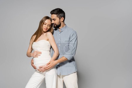 Trendy and bearded man hugging fair haired and pregnant woman while touching belly while standing together on grey background, expecting parents concept, baby bump, husband and wife 