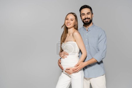 Smiling and bearded man hugging belly of fashionable pregnant woman and looking at camera while standing together isolated on grey, expecting parents concept, baby bump, husband and wife 