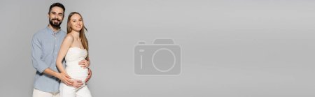 Photo for Smiling and bearded man hugging fashionable pregnant woman and looking at camera while standing isolated on grey with copy space, expecting parents concept, banner, baby bump, husband and wife - Royalty Free Image