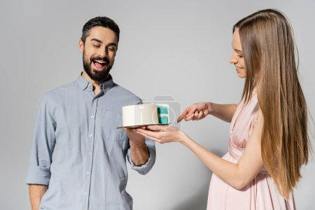 Excited and bearded man holding cake near cheerful pregnant wife with knife during baby shower party on grey background, expecting parents concept, gender party, it`s a boy