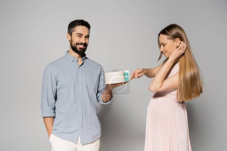 Positive and bearded man holding cake near elegant pregnant wife in pink dress during gender party on grey background, expecting parents concept, gender party, it`s a boy, cutting cake 