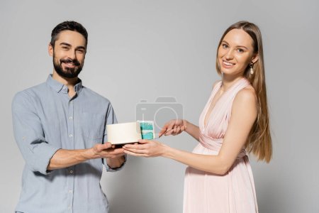 Cheerful and stylish couple looking at camera while cutting blue cake during baby shower celebration on grey background, expecting parents concept, gender party, it`s a boy