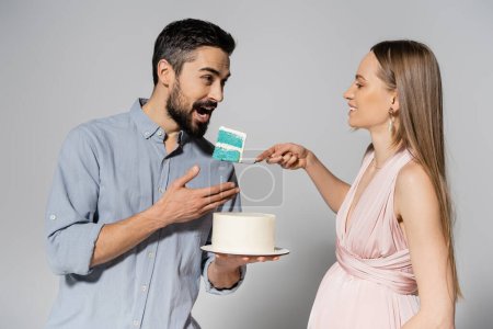 Femme enceinte souriante en robe rose tenant le gâteau bleu près du mari avec la bouche ouverte pendant la fête de genre sur fond gris, attendant concept de parents, c'est un garçon 