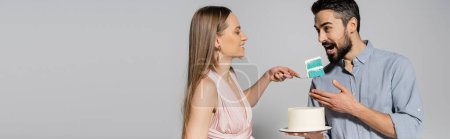 Cheerful and elegant woman holding blue cake near husband opening mouth during gender party celebration on grey background, expecting parents concept, it`s a boy, banner 