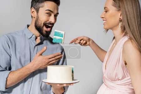 Smiling and elegant pregnant woman feeding husband with blue cake during gender party celebration isolated on grey, expecting parents concept happiness, it`s a boy