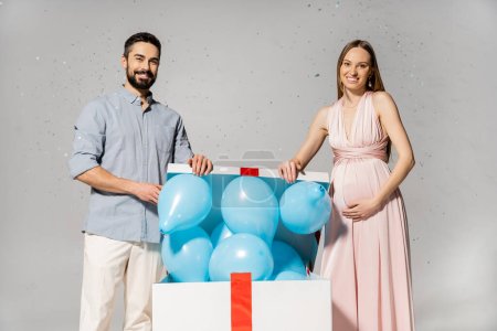 Positive pregnant woman in elegant dress opening bog gift box with blue balloons while standing near husband under confetti during baby shower on grey background, gender party, it`s a boy