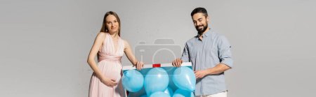 Elegante und schwangere Frau öffnet Geschenkbox mit blauen Luftballons neben fröhlichem Ehemann und Konfetti während Baby-Shower-Feier auf grauem Hintergrund, Geschlechterparty, es ist ein Junge, Banner 
