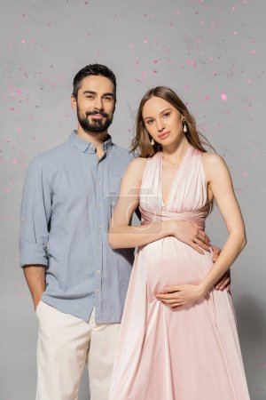 Portrait of smiling man hugging elegant and pregnant wife in pink dress while standing under confetti during baby shower party on grey background, expecting parents concept 
